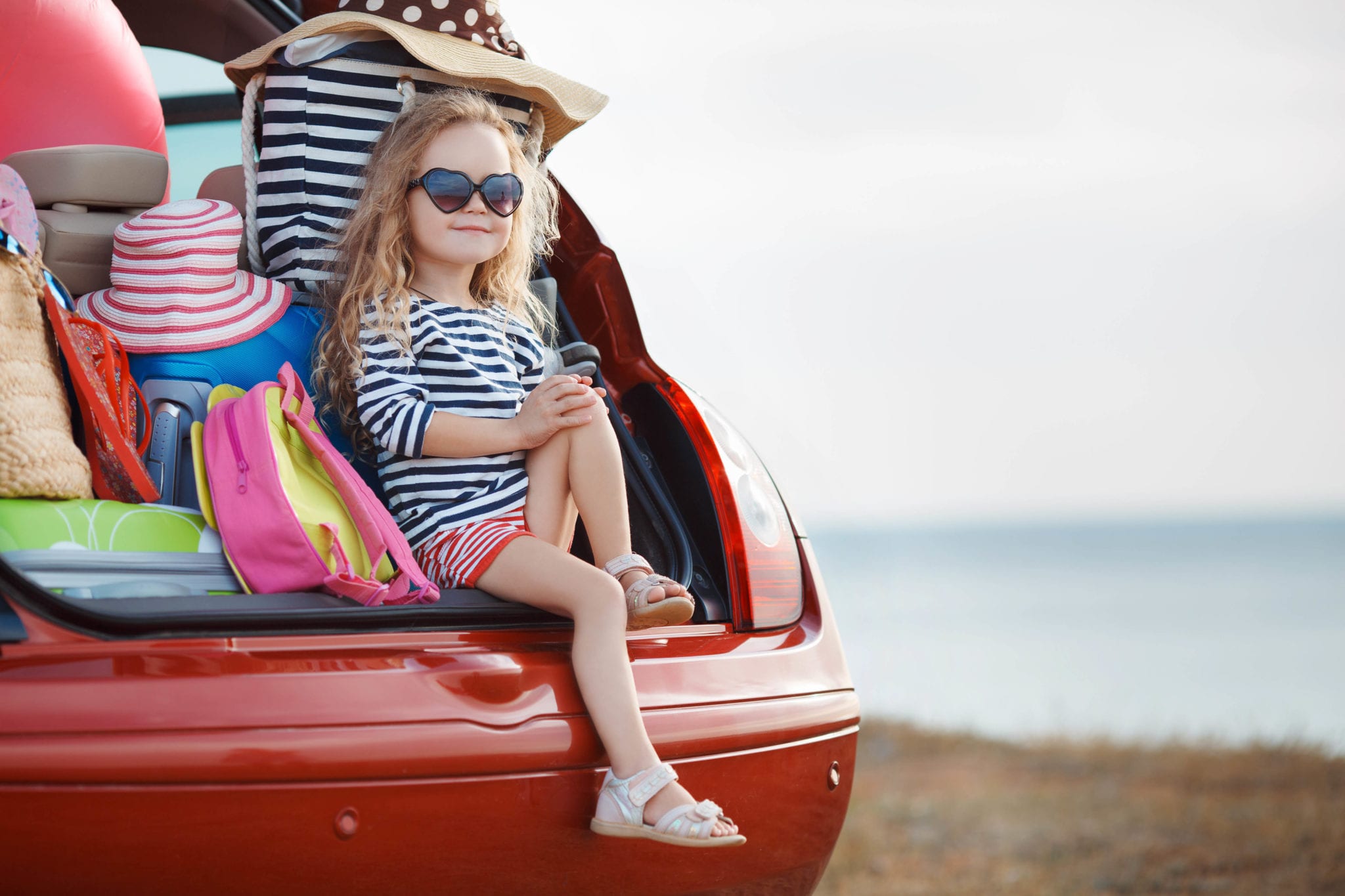 Young girl sitting in the open boot of a car with luggage and smiling